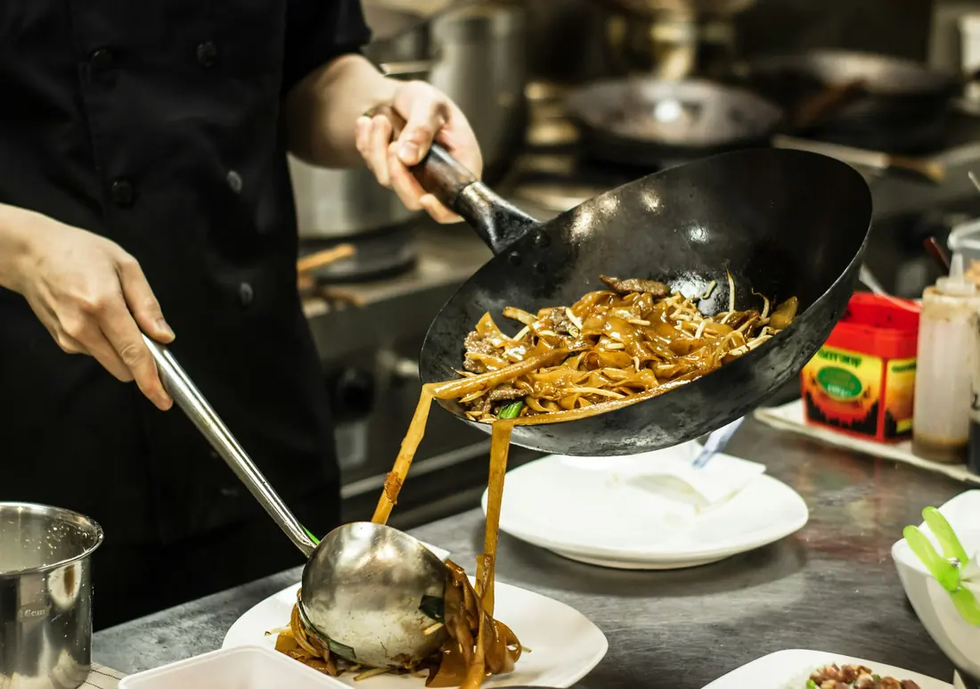 A cook tossing noodles in a wok with tongs