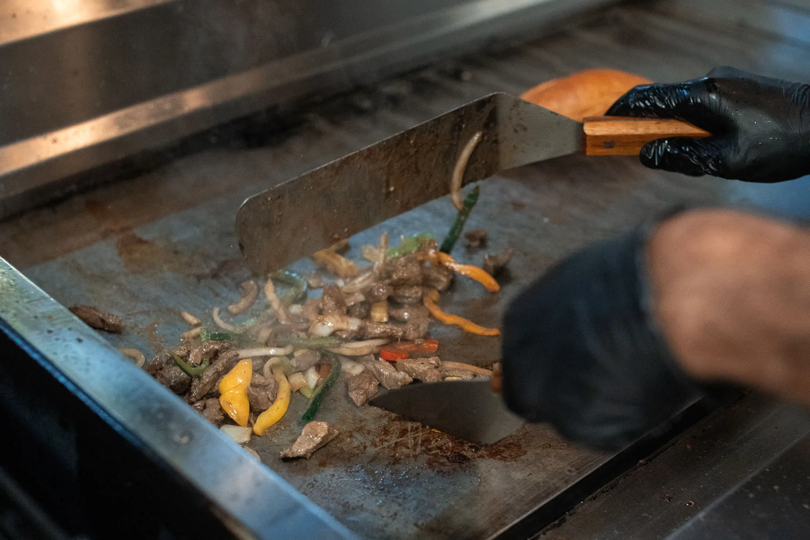Meat, onions and peppers being turned with a spatula on a flat top grill