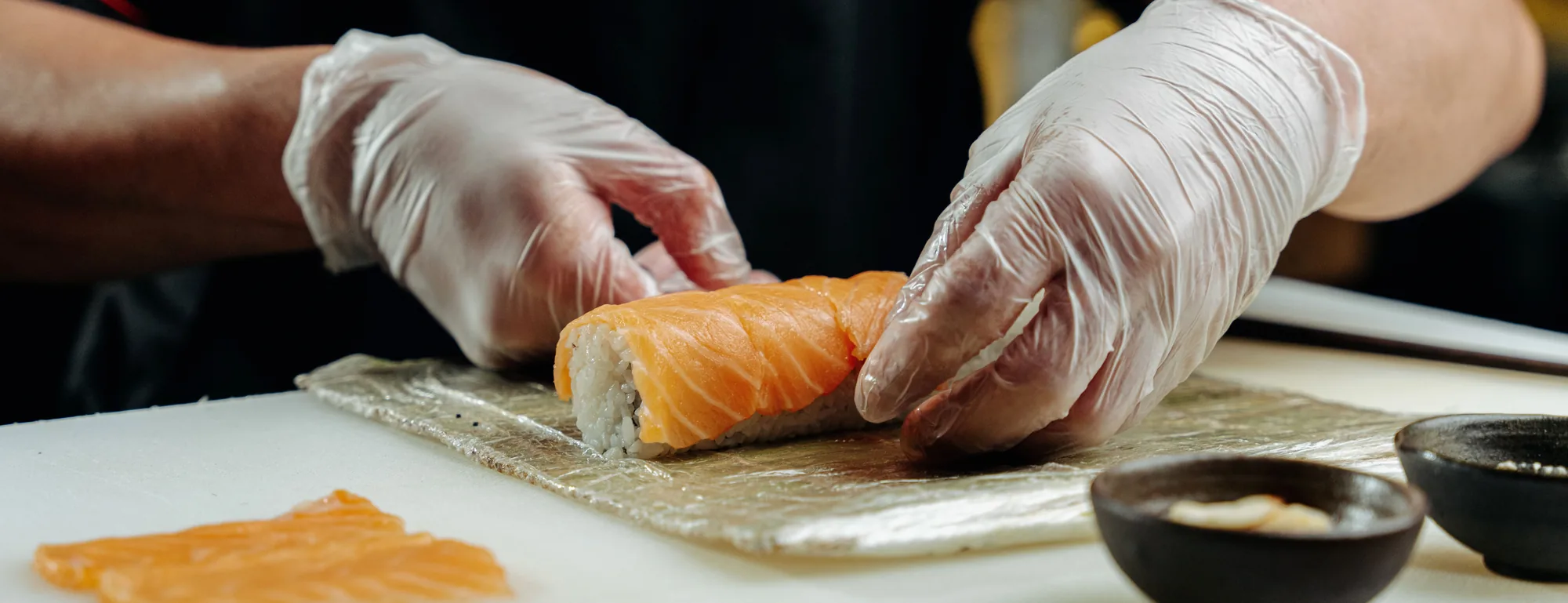 Gloved hands finishing a salmon roll on a bamboo rolling mat